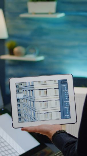 Architect holding digital tablet with building model for construction layout in office. Man engineer working on blueprint plan with touch screen monitor and device for architectural project